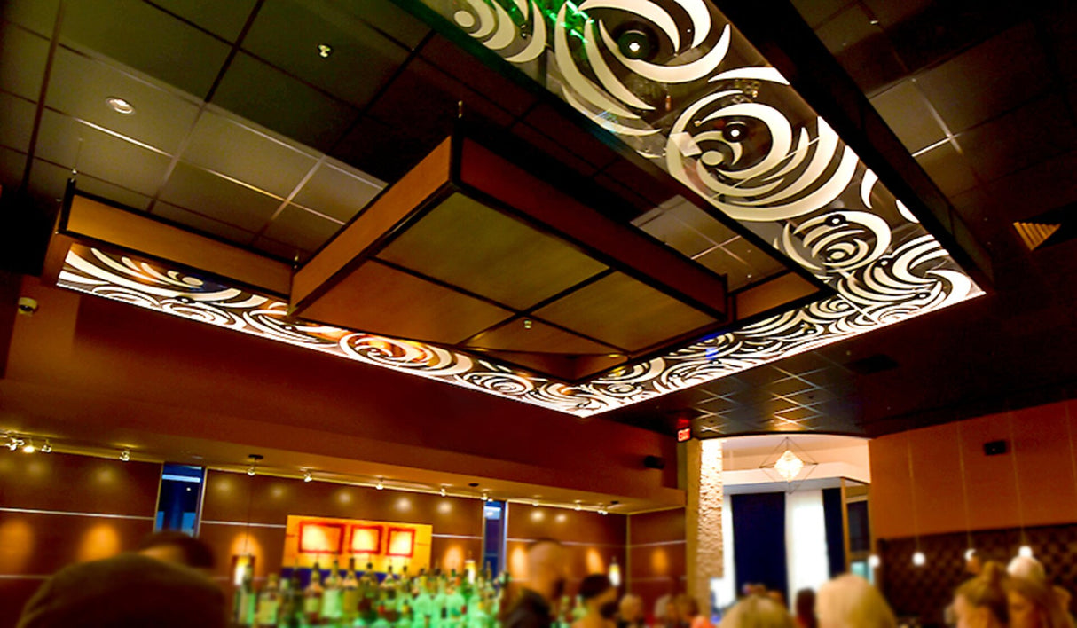 A stylish restaurant features a decorative ceiling with swirling patterns illuminated by the Klus KRAV-56 Edge-lit LED Extrusion for 1/4 glass panels, warm lighting, and a vibrant bar area where people gather, creating an inviting atmosphere.