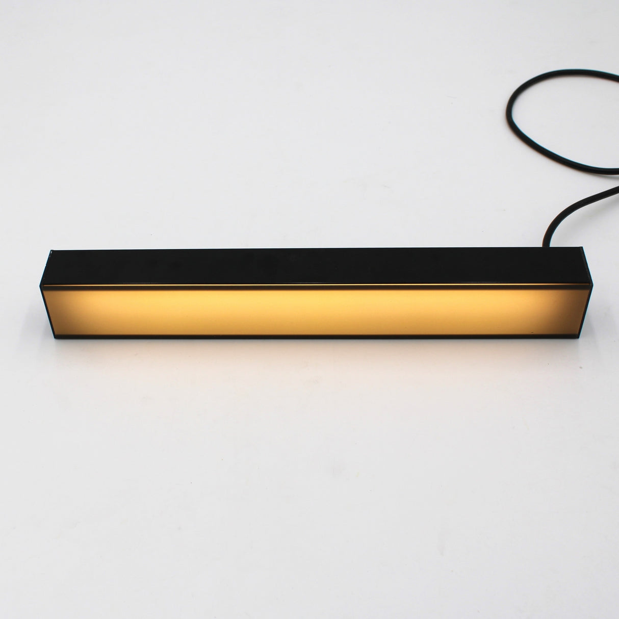 The 2 Wide Aluminum Linear LED Lighting Profile (Model PL55-FL) in black emits a warm yellow glow onto a white surface, with a black power cord extending from its right side.