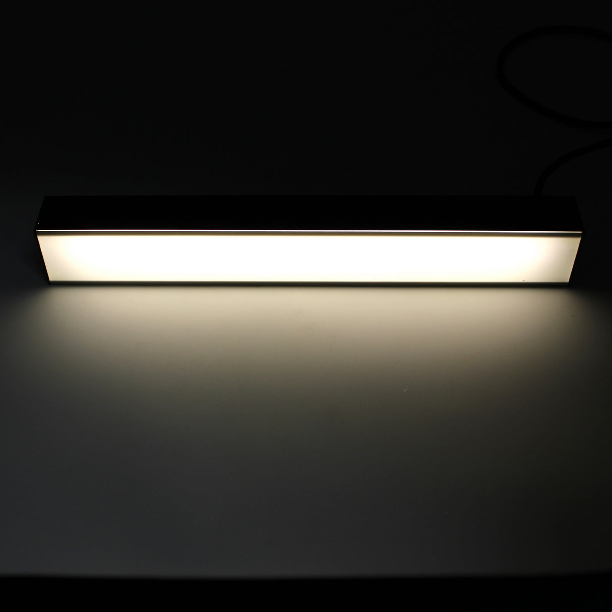 The 2 Wide Aluminum Linear LED Lighting Profile (Model PL55-FL) emits a bright, even glow across a nearby surface in a dark room, with its sleek aluminum design and intense illumination standing out against the mostly black background.