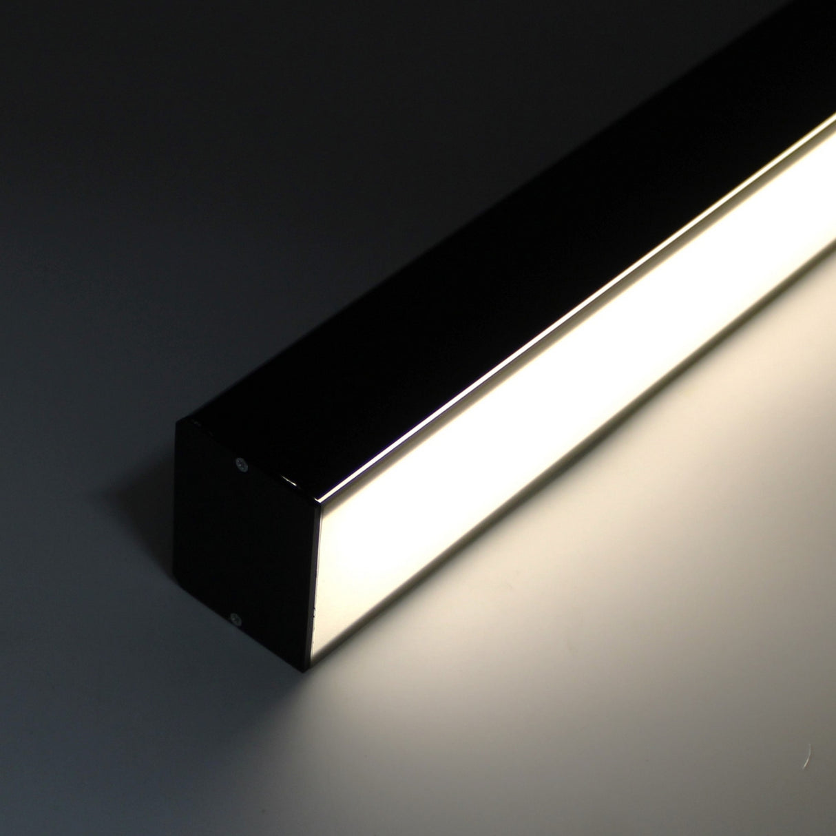 A close-up of the 2 Wide Aluminum Linear LED Lighting Profile (Model PL55-FL) showcases its sleek black housing and sharp edges, emitting a bright white glow as it rests on a dark surface.