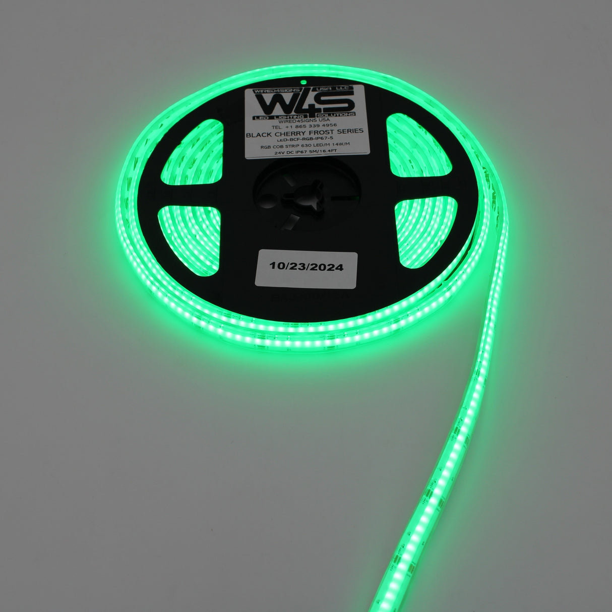 A spool of RGB COB IP67 LED Strip (24V) ~ Black Cherry Frost Series glows green on a white surface, with a label and a sticker showing the date 10/23/2024.