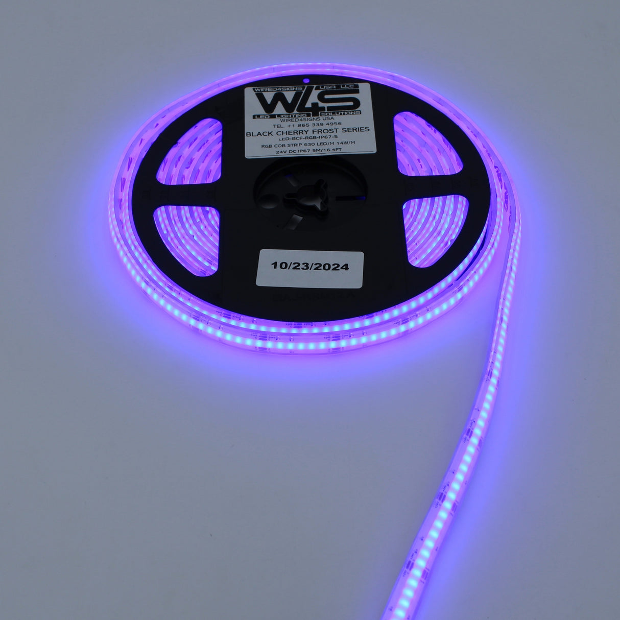 A reel of WS RGB COB IP67 LED Strip (24V) ~ Black Cherry Frost Series emits a purple glow. The labeled reel, dated 10/23/2024, sits on a white surface with the strip extending outward.