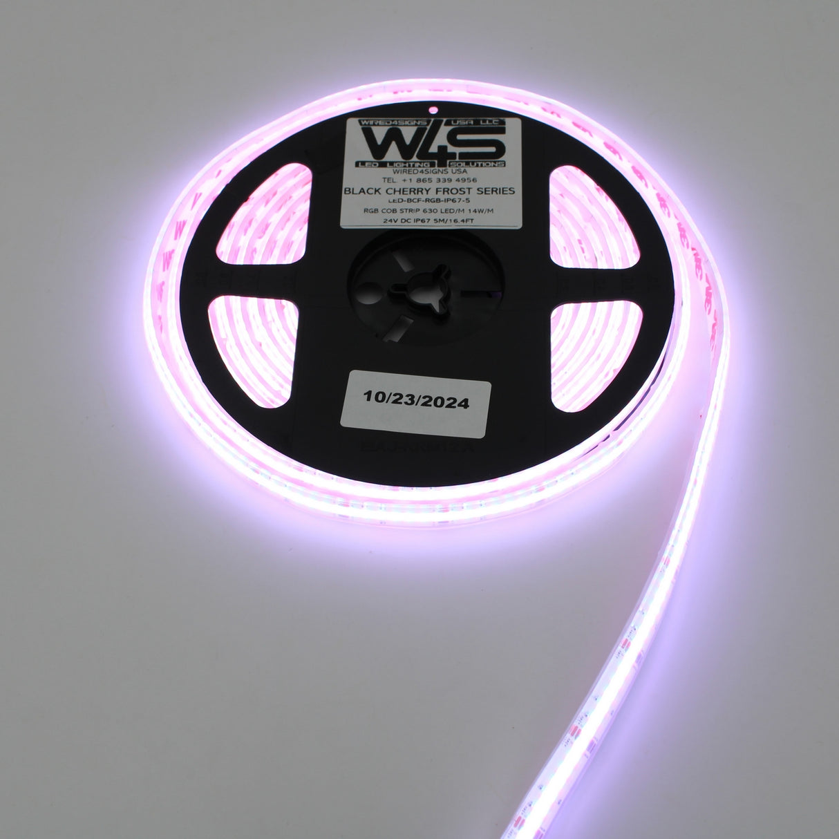 A reel of RGB COB IP67 LED Strip (24V) from the Black Cherry Frost Series emits a vibrant pinkish-purple glow, partially unrolled on a white surface with a label and date sticker marked “10/23/2024.”.