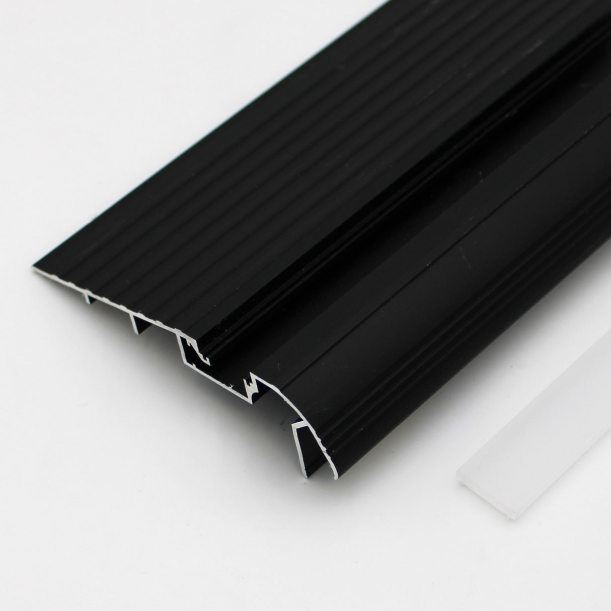 Close-up of the LED Diffuser for Treadable Easy-On Profiles placed next to a black, grooved metal profile with a geometric cross-section on a white surface.