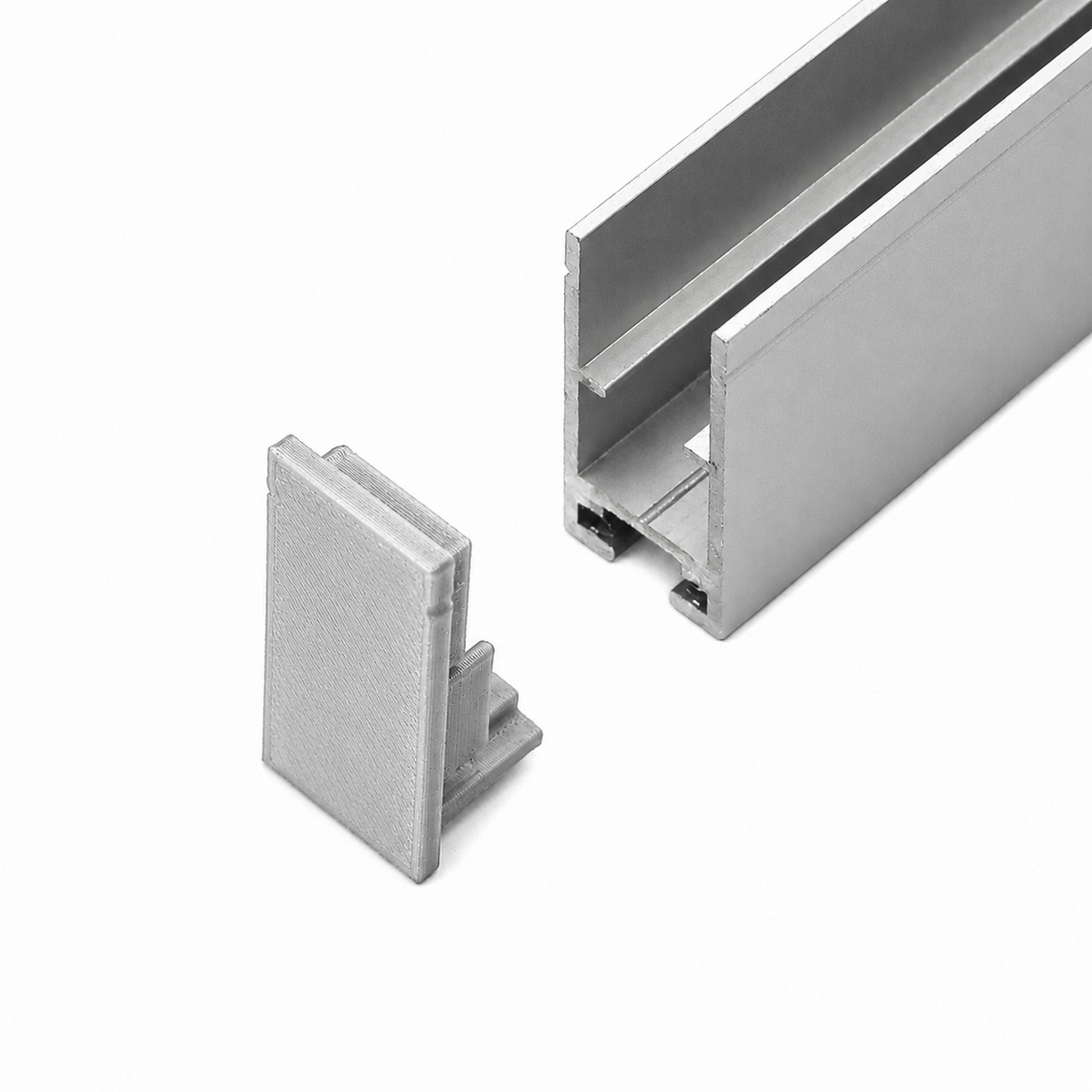 A silver metal rectangular Hannover Profile channel with an open groove is shown next to a Plastic End Cap for Hannover Profile, both placed on a white background. The End Cap appears designed to fit into the open end of the channel.