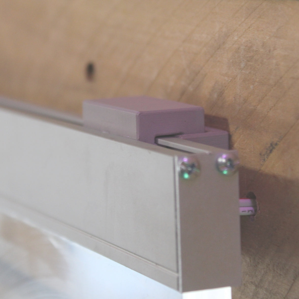 Close-up of the Wall Mounting Bracket for Hannover Profile secured with screws to a wooden surface, holding the edge of a rectangular panel—perfect for mounting edge-lit acrylic signage in contemporary displays.
