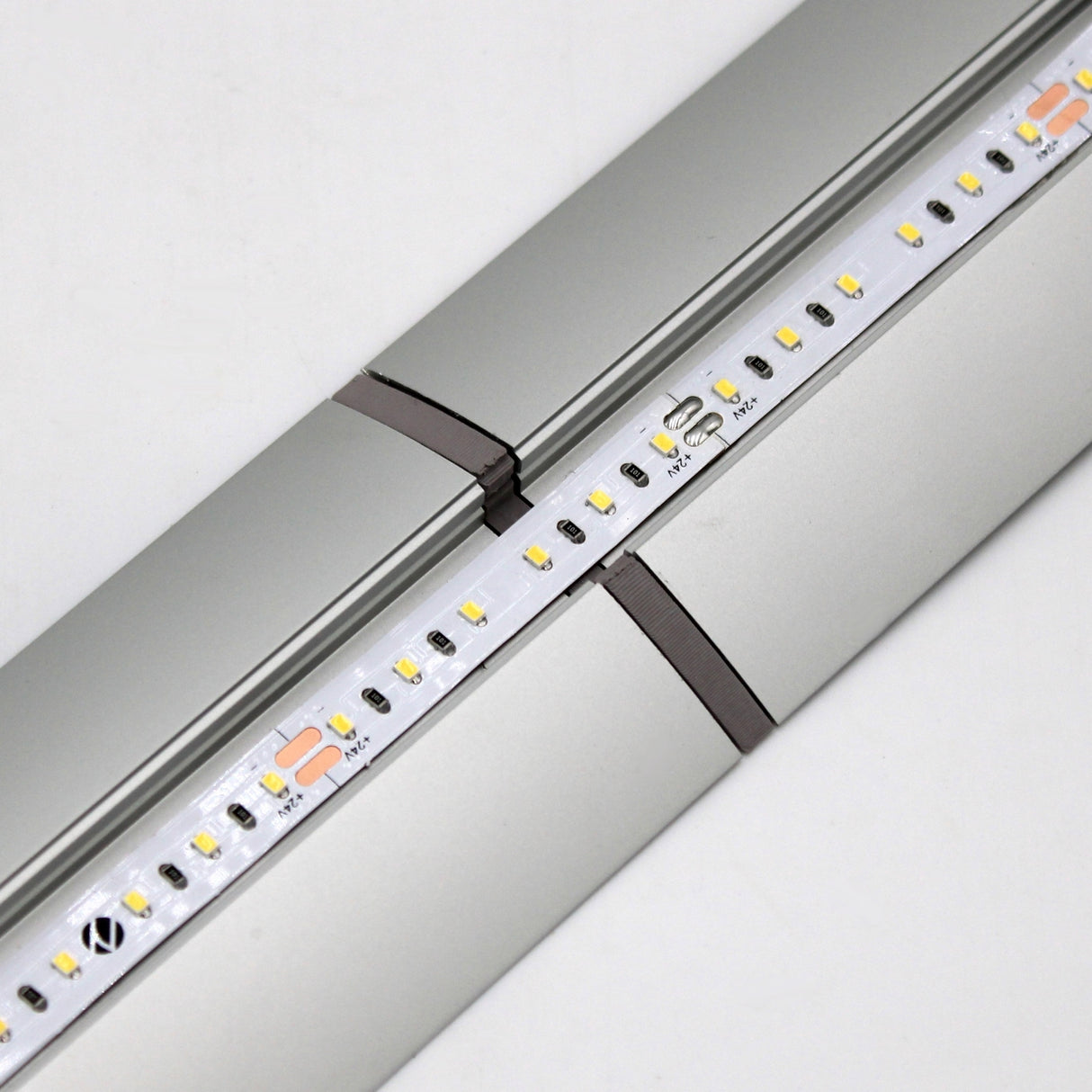 Close-up of a silver LED strip light on an Alu-Flat profile, featuring a seamless connection between two sections using the Plastic Straight Connector for Alu-Flat Profile. Small yellow and white LEDs are visible on the fixture.