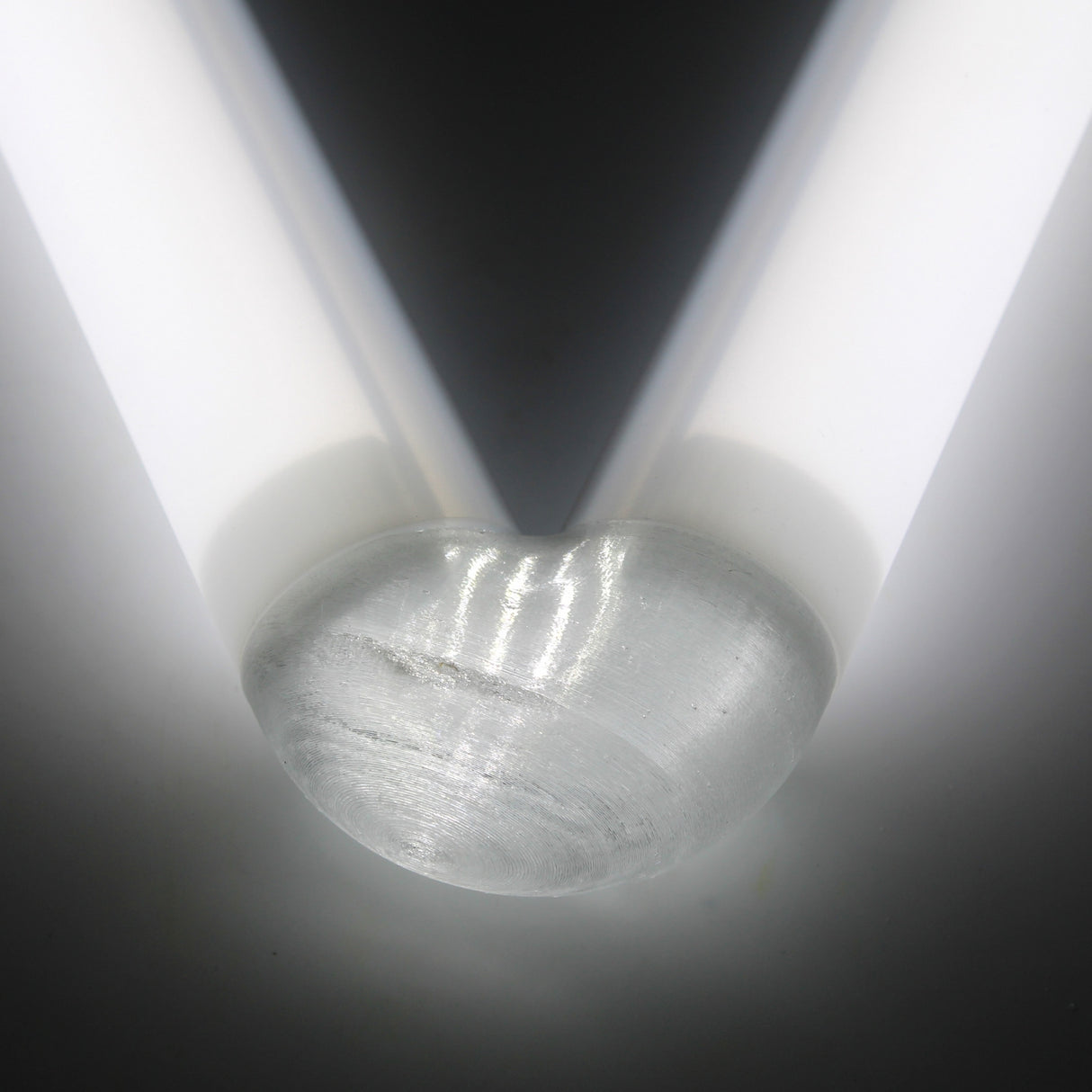 A close-up of the rounded end of a Translucent Elbow for Oslo Profile placed between two parallel white light tubes, forming a V-shaped pattern of light and shadow on a neutral background.
