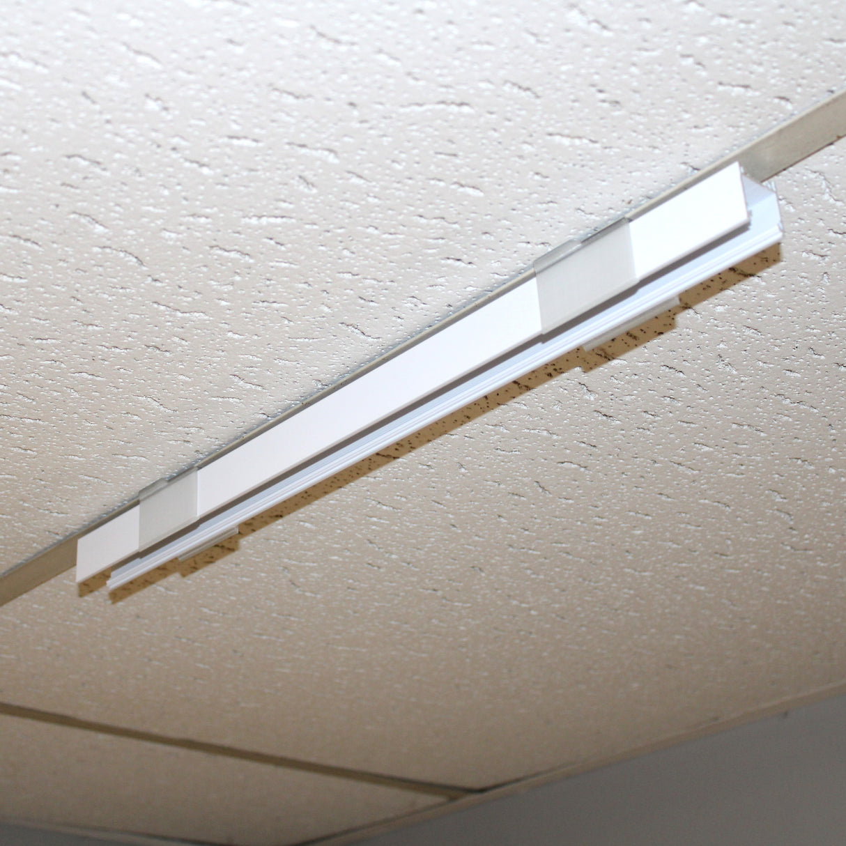 A Drop Ceiling T-Bar Linea20 Clip is installed in a white, textured ceiling with acoustic tiles and a drop-ceiling frame. The light fixture is currently turned off.
