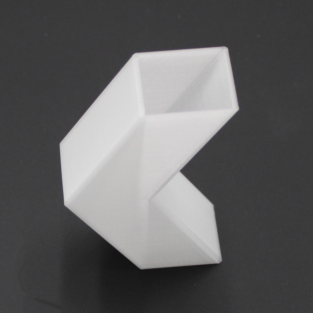 A translucent, angular 3D-printed Translucent Elbow for Visby Profile with a zigzag shape and three connected rectangular segments is shown on a dark gray background.