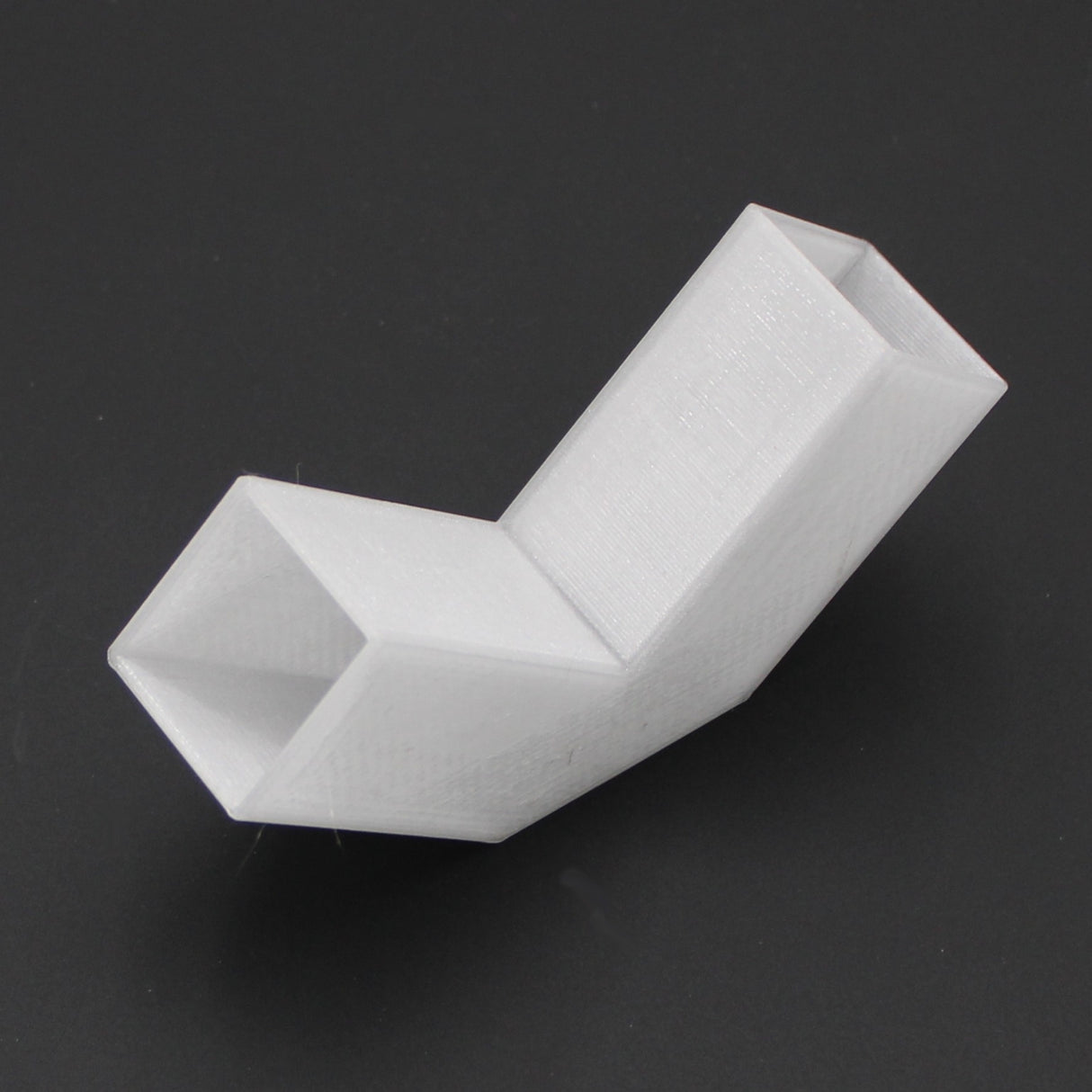 A Translucent Elbow for Visby Profile, featuring a white, 3D-printed L-shaped design with two rectangular openings and an angled section, is displayed against a black background.