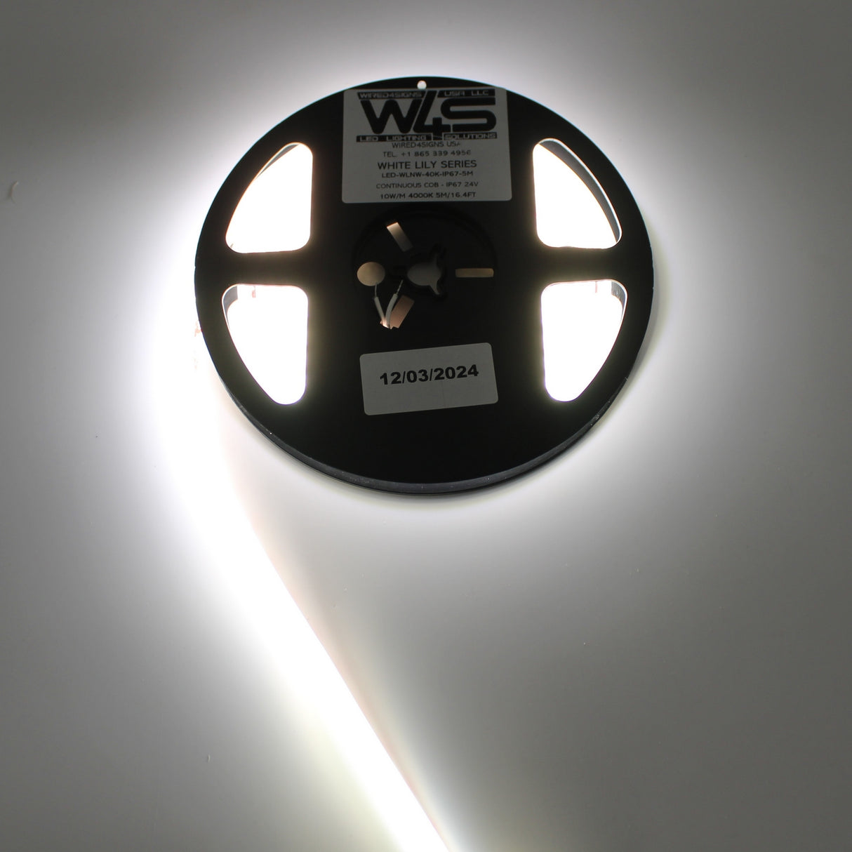A spool of Continuous COB IP67 LED Strip (24V) ~ White Lily Series emits bright, dot-free light in a circular glow from its attached strip. A 12/03/2024 date sticker is on the center—ideal for stylish, decorative lighting.