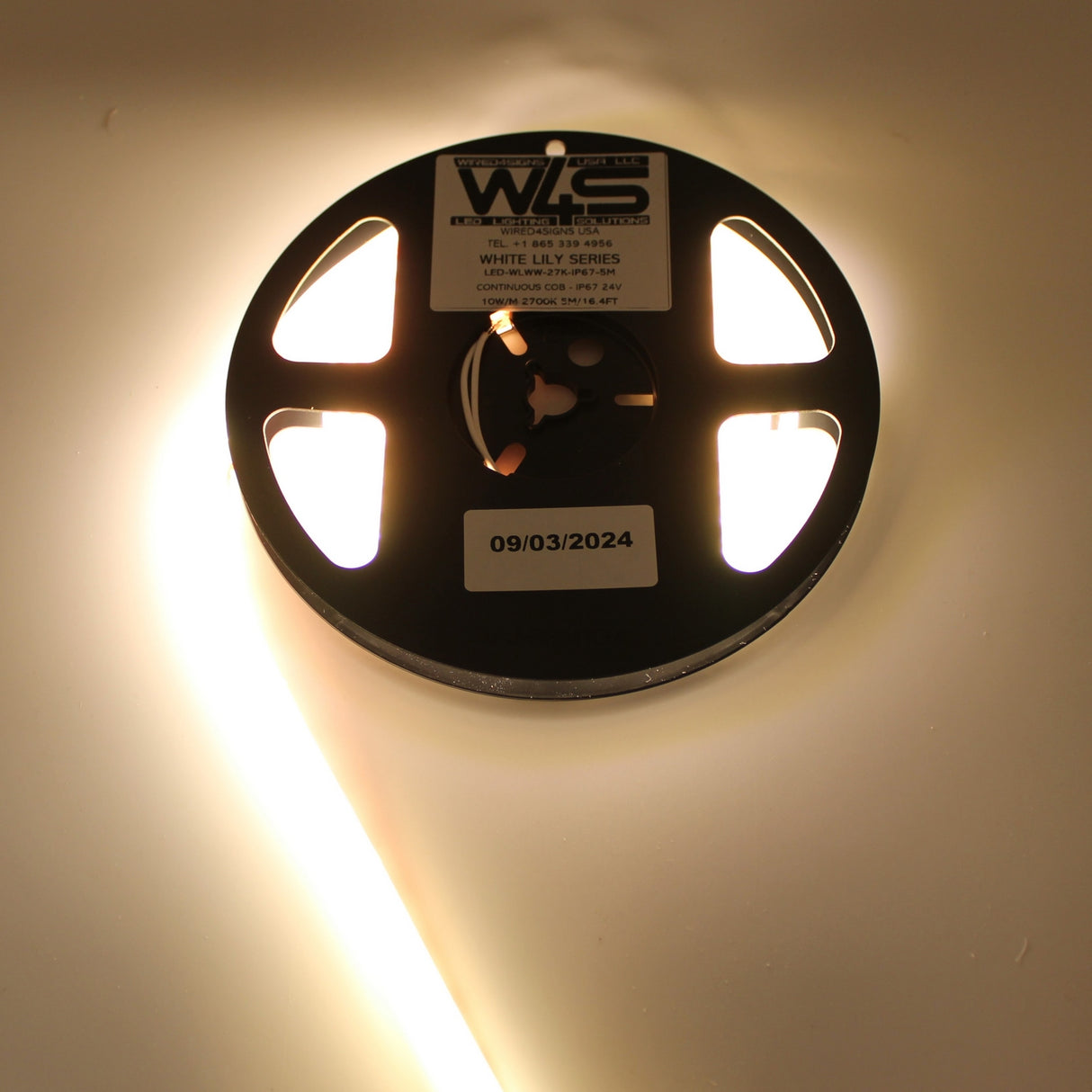 A partially unspooled Continuous COB IP67 LED Strip (24V) ~ White Lily Series glows brightly on a white surface, with a reel labeled “White Lily Series” and a “09/03/2024” sticker—ideal for dot-free decorative lighting.