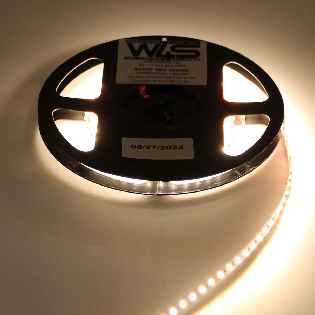 A reel of White High CRI IP20 LED Strip (24V) ~ White Iris Series glows warmly on a flat surface, emitting white light from the unspooled strip. The reel is labeled WHITE IRIS SERIES with a date sticker 08/27/2024.
