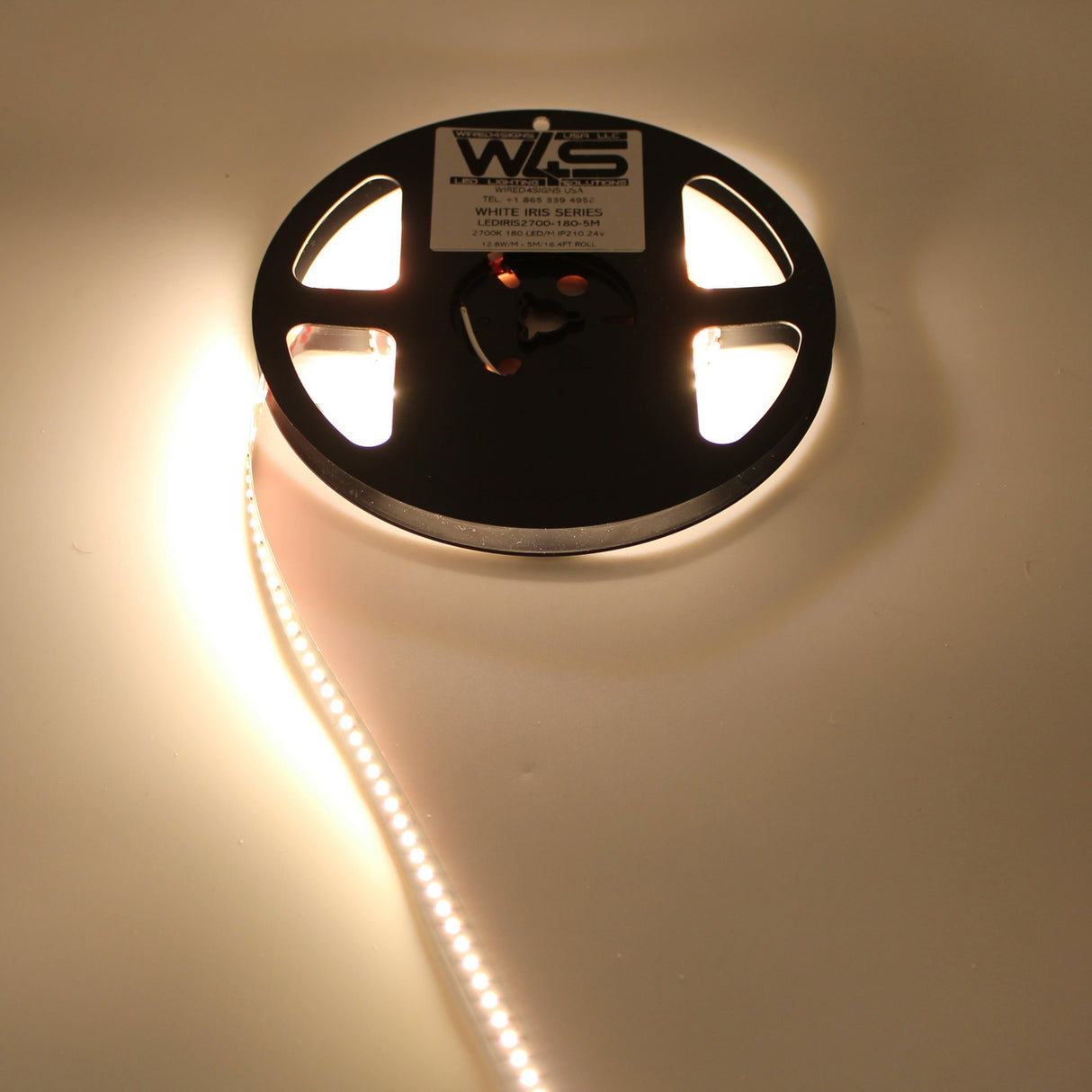A partially unspooled reel of White High CRI IP20 LED Strip (24V) ~ White Iris Series glows warmly on a white surface, emitting a rich warm white light. The reel features a label on top.