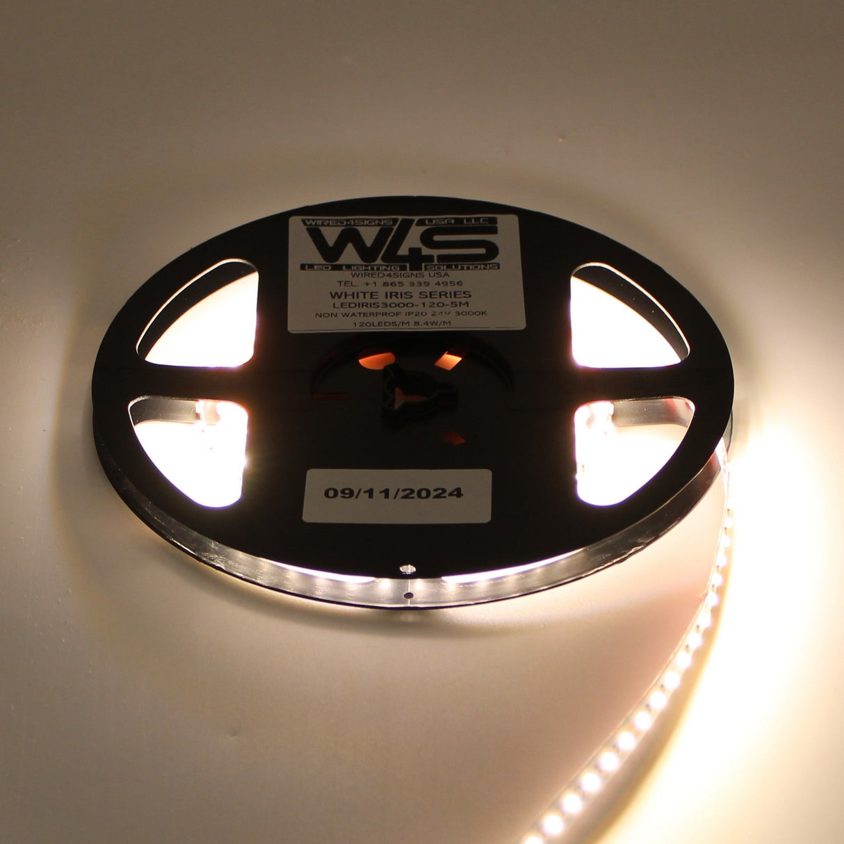 A reel of White High CRI IP20 LED Strip (24V) ~ White Iris Series emits a warm white glow. Its label displays text and the date 09/11/2024. The partially unspooled, lit strip highlights quality color temperatures.