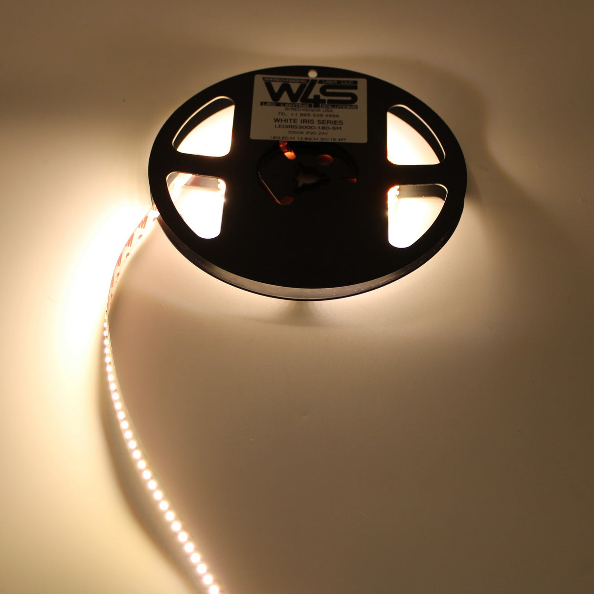 A White High CRI IP20 LED Strip (24V) ~ White Iris Series is partially unspooled and glowing brightly on a smooth, light surface, forming a curved line away from the black reel. The strip offers various color temperatures.