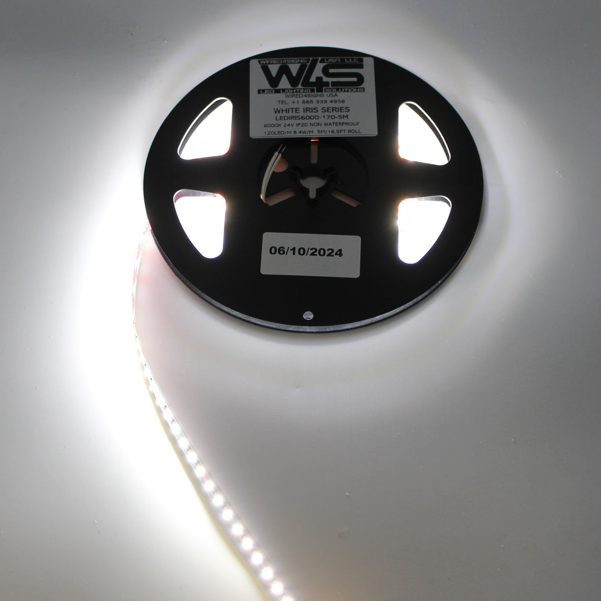 A black reel labeled White Iris Series holds a partially unspooled 24V White High CRI IP20 LED strip with bright white LEDs, lying on a white surface. A sticker on the reel shows the date 06/10/2024.