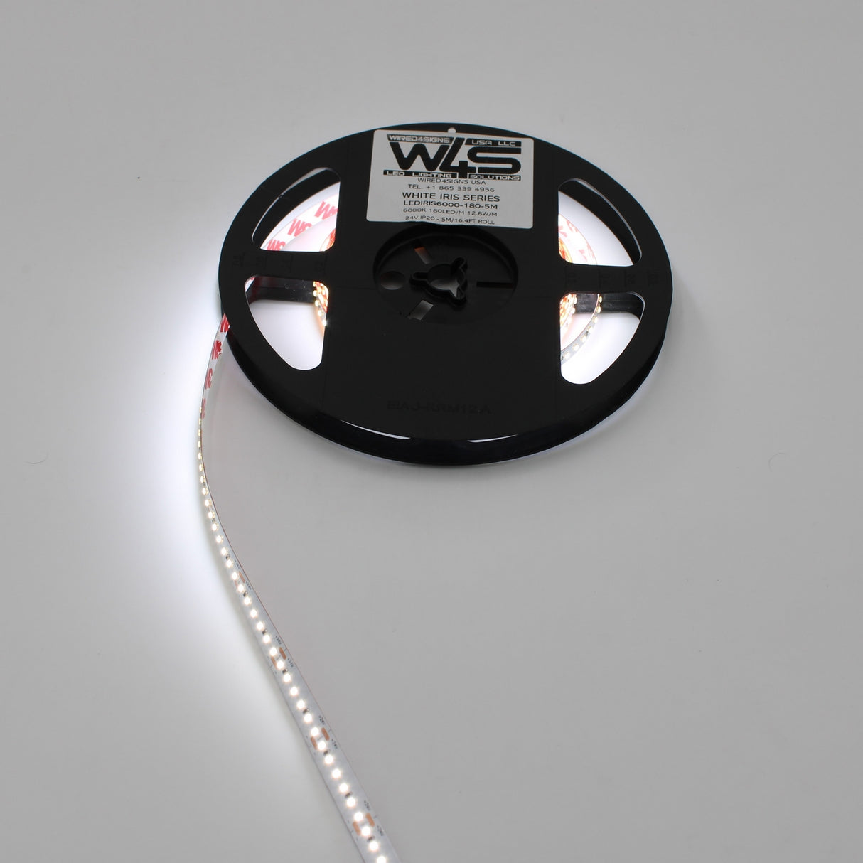 A White High CRI IP20 LED Strip (24V) ~ White Iris Series reel is partially unspooled on a plain white surface, emitting bright light. The labeled reel showcases quality branding, ideal for versatile color temperatures.