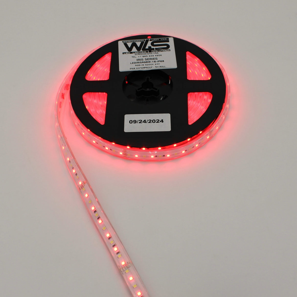 A reel of High CRI RGBW IP68 Waterproof LED Strip (24V) ~ Iris Series glows red, partially unspooled on a white surface. The reel’s label displays product and date info.