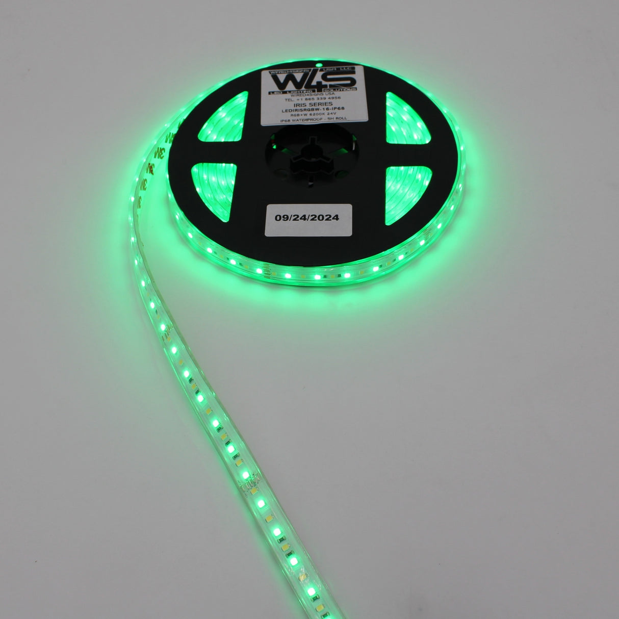 A reel of High CRI RGBW IP68 Waterproof LED Strip (24V) ~ Iris Series glows green, partially unraveled on a white background. The reels label displays text and the date 09/24/2024.