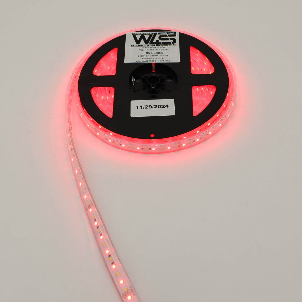 A spool of High CRI RGBW IP68 Waterproof LED Strip (24V) ~ Iris Series, partially unrolled and glowing red, rests on a white surface. A label with text and the date 11/29/2024 is visible on the reel.