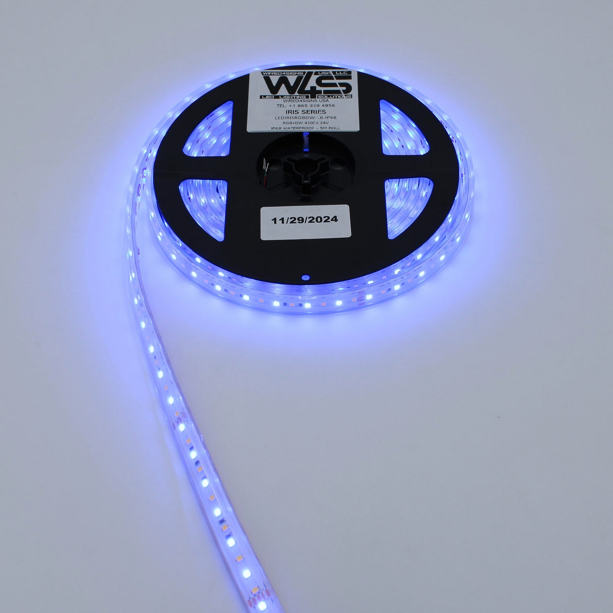 A spool of High CRI RGBW IP68 Waterproof LED Strip (24V) ~ Iris Series is partially unrolled on a white surface, illuminated, with a label dated 11/29/2024 visible on the spool.