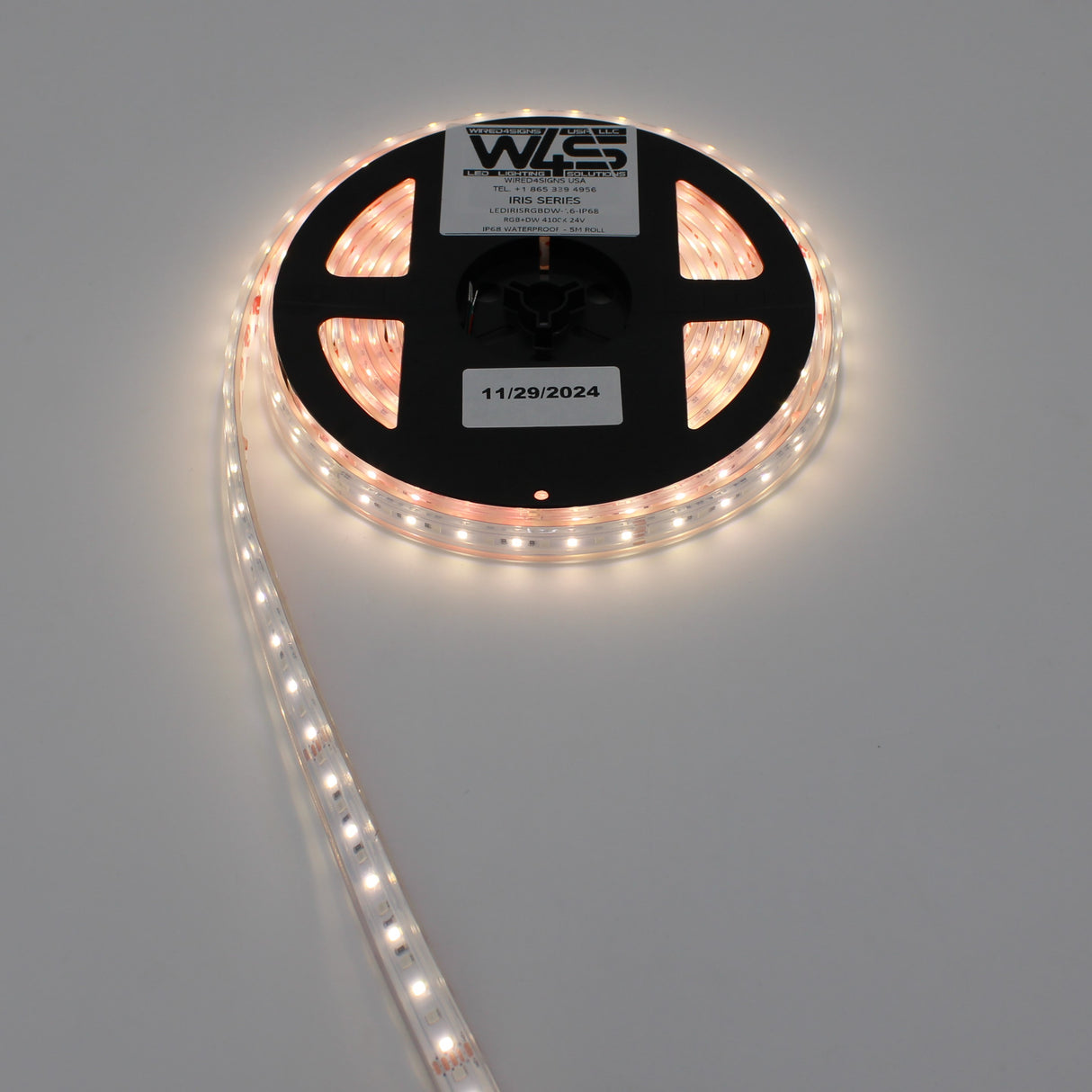 A coiled reel of High CRI RGBW IP68 Waterproof LED Strip (24V) ~ Iris Series is partially unspooled and glowing warm white. The label displays the brand WAS and an expiration date of 11/29/2024.