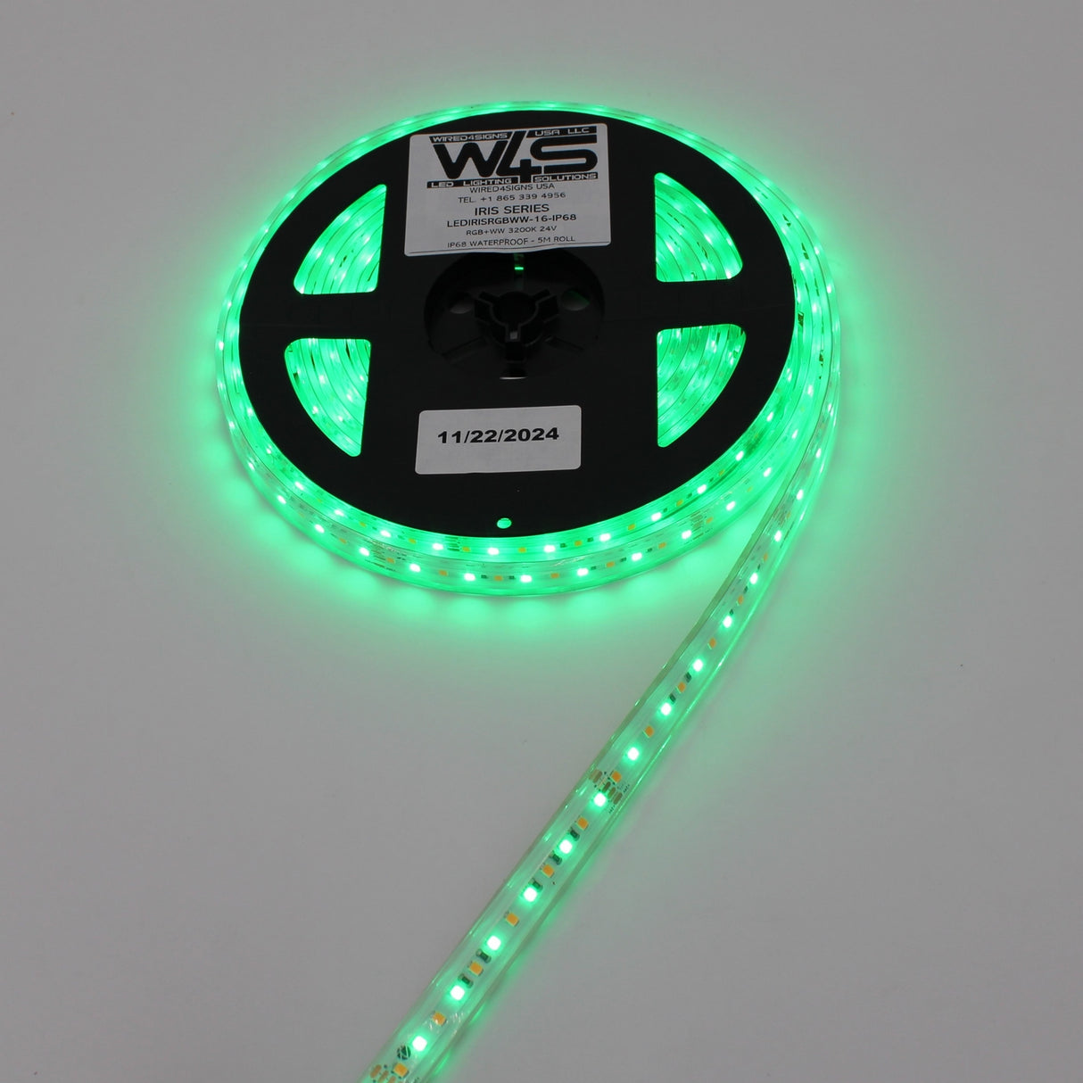 A spool of High CRI RGBW IP68 Waterproof LED Strip (24V) ~ Iris Series glows green, partially unspooled on a plain background. The reel displays a label with WLS and the date 11/22/2024.