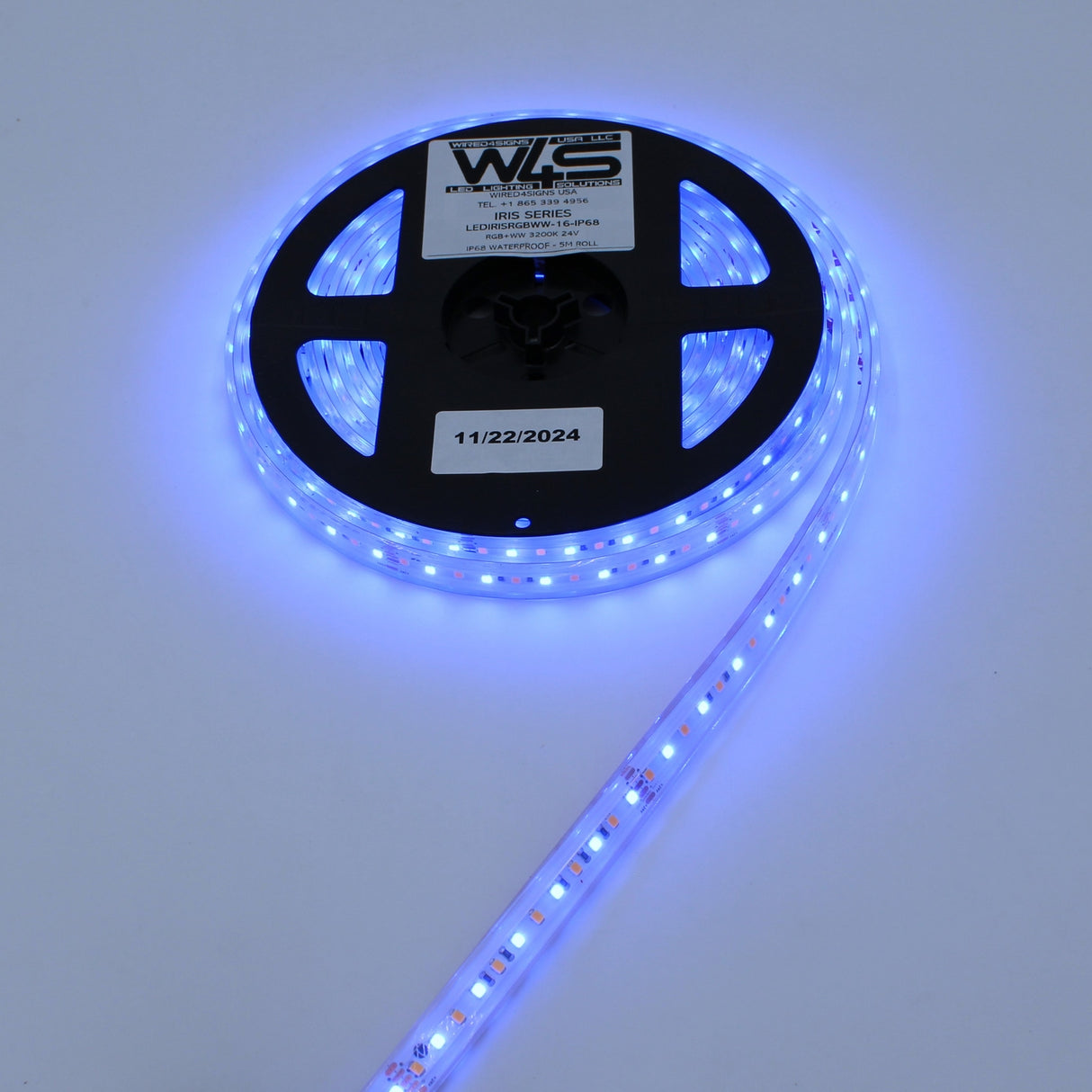 A spool of High CRI RGBW IP68 Waterproof LED Strip (24V) ~ Iris Series is partially unrolled on a white surface, glowing brightly. A label marked 11/22/2024 can be seen on the reel.