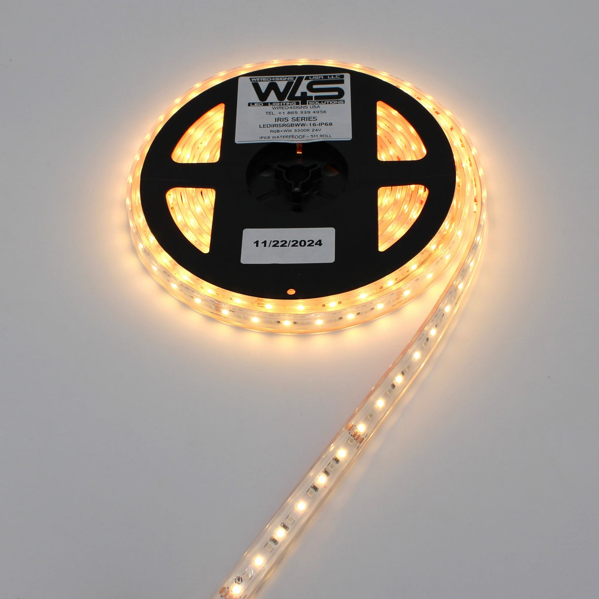 A spool of High CRI RGBW IP68 Waterproof LED Strip (24V) ~ Iris Series glows while partially uncoiled on a white surface. The black reel displays product labels, including a date marked 11/22/2024.