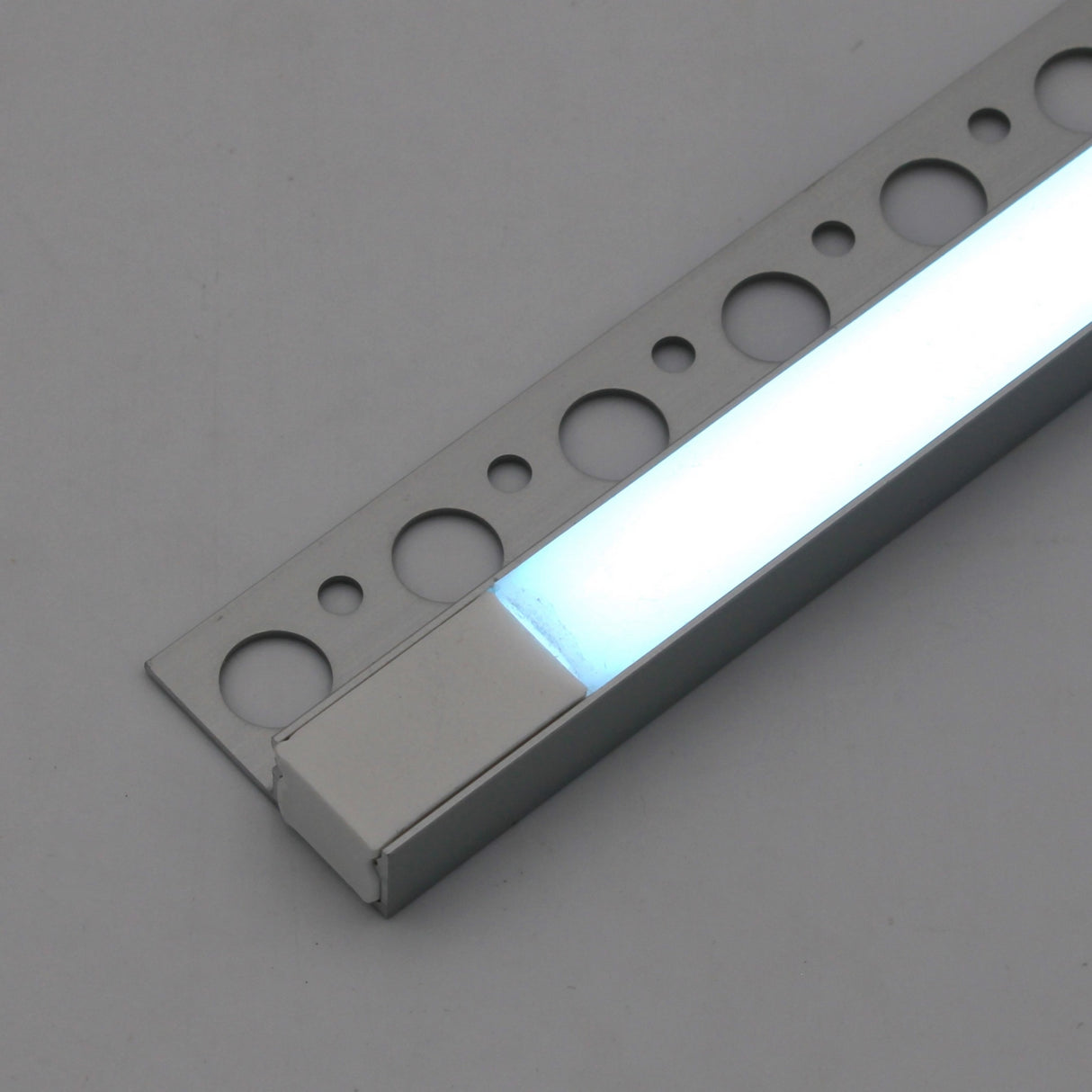 A close-up of the RGB LED Tile Edge Light Strip Kit (24V) ~ Nicho Series, a rectangular aluminum channel with a silver frame and circular cutouts, placed on a gray surface. Its RGB LEDs emit a soft white glow.