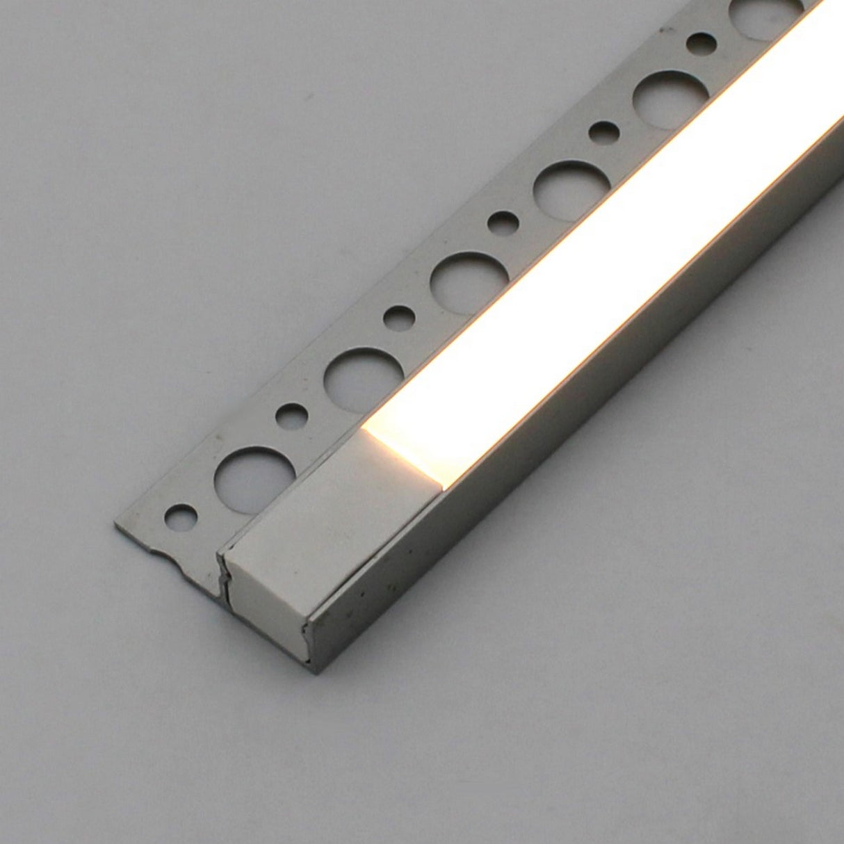 A close-up of the White LED Tile Edge Light Strip Kit (24V) ~ Nicho Series, recessed in an aluminum channel with round perforations, shown against a plain light gray background.