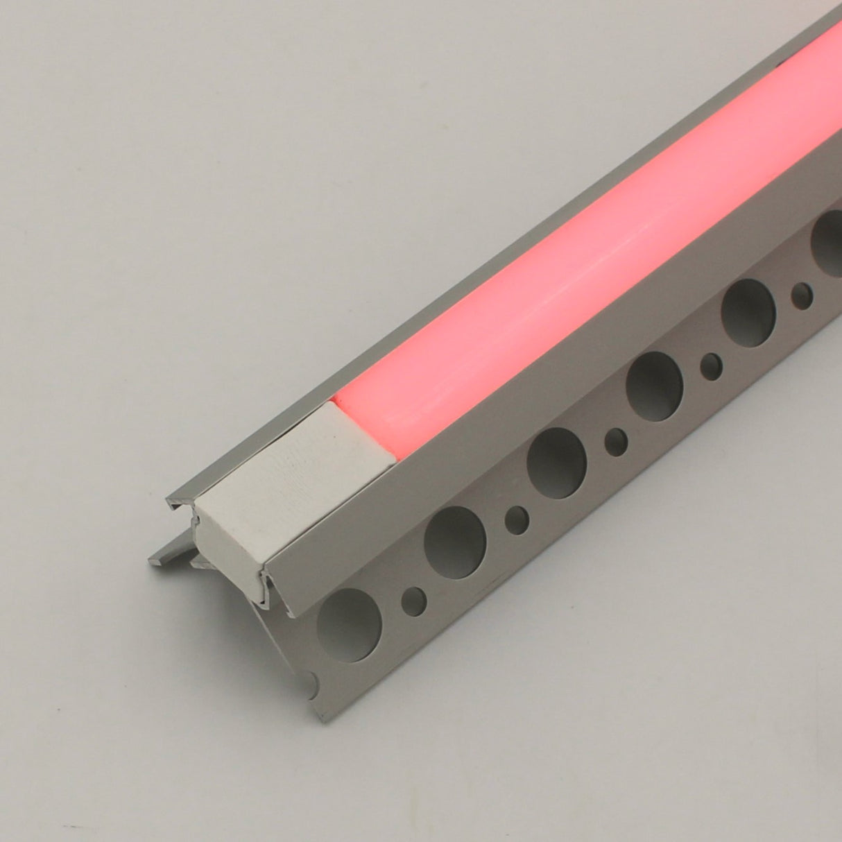 A close-up of the RGB LED Tile Light Strip Kit for Outside Corners (24V) ~ Nicho Series, featuring an aluminum channel with a frosted cover emitting soft pink light, suitable for recessed or surface mounting, displayed on a light gray surface.