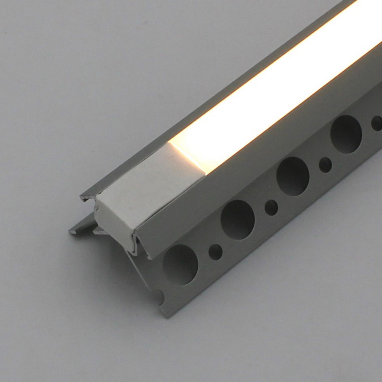 A close-up of the Nicho Series White LED Tile Light Strip Kit for Outside Corners (24V) installed in an aluminum channel with circular holes, casting a warm white glow against a plain background.
