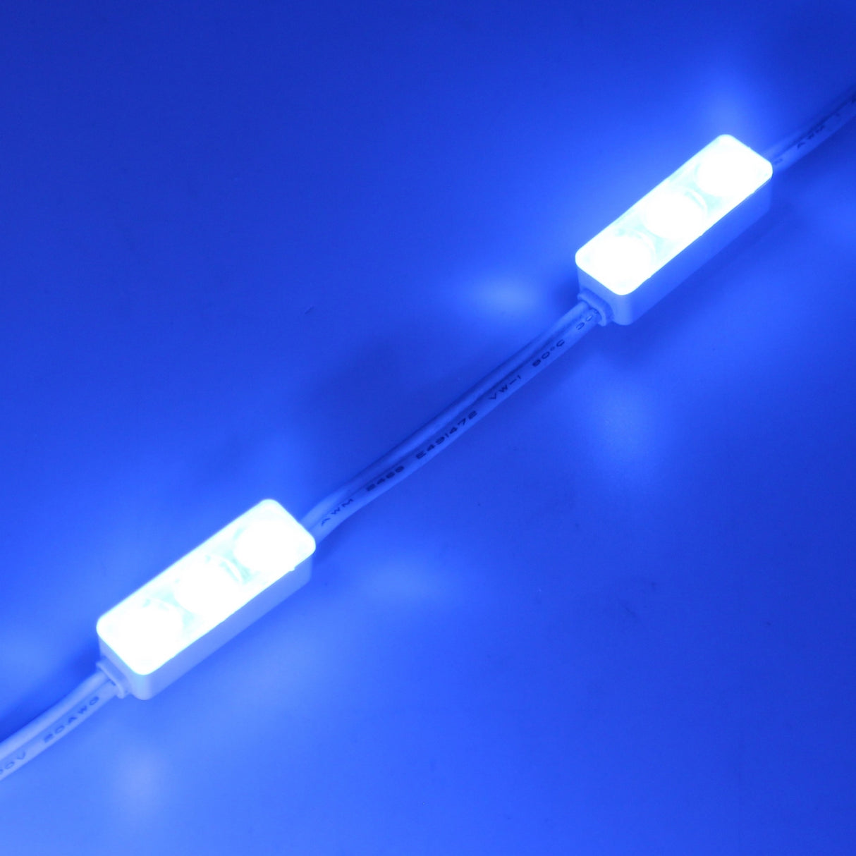Two Mini3 Color 160 Mini LED Module ~ Wide Angle Series units, linked by a white wire, emit a bright blue glow against a blue background, captured with a wide-angle lens.