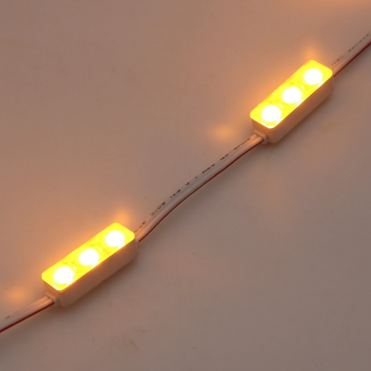 Two Mini3 Color 160 Mini LED Module ~ Wide Angle Series fixtures, each with three yellow LEDs, are joined by wires and cast a warm glow on a neutral surface—ideal for showcasing the illumination of these waterproof IP68-rated modules.