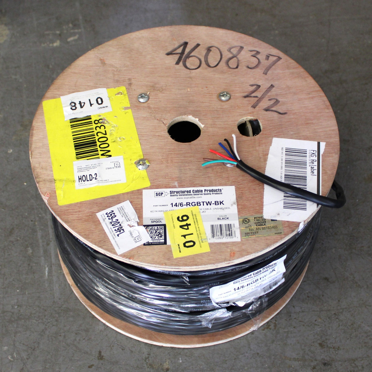 A large spool of RGB+TW 6-Core PVC Coated Wire, black with multiple exposed colored cores and several yellow and white labels and barcodes, rests on a gray concrete floor.