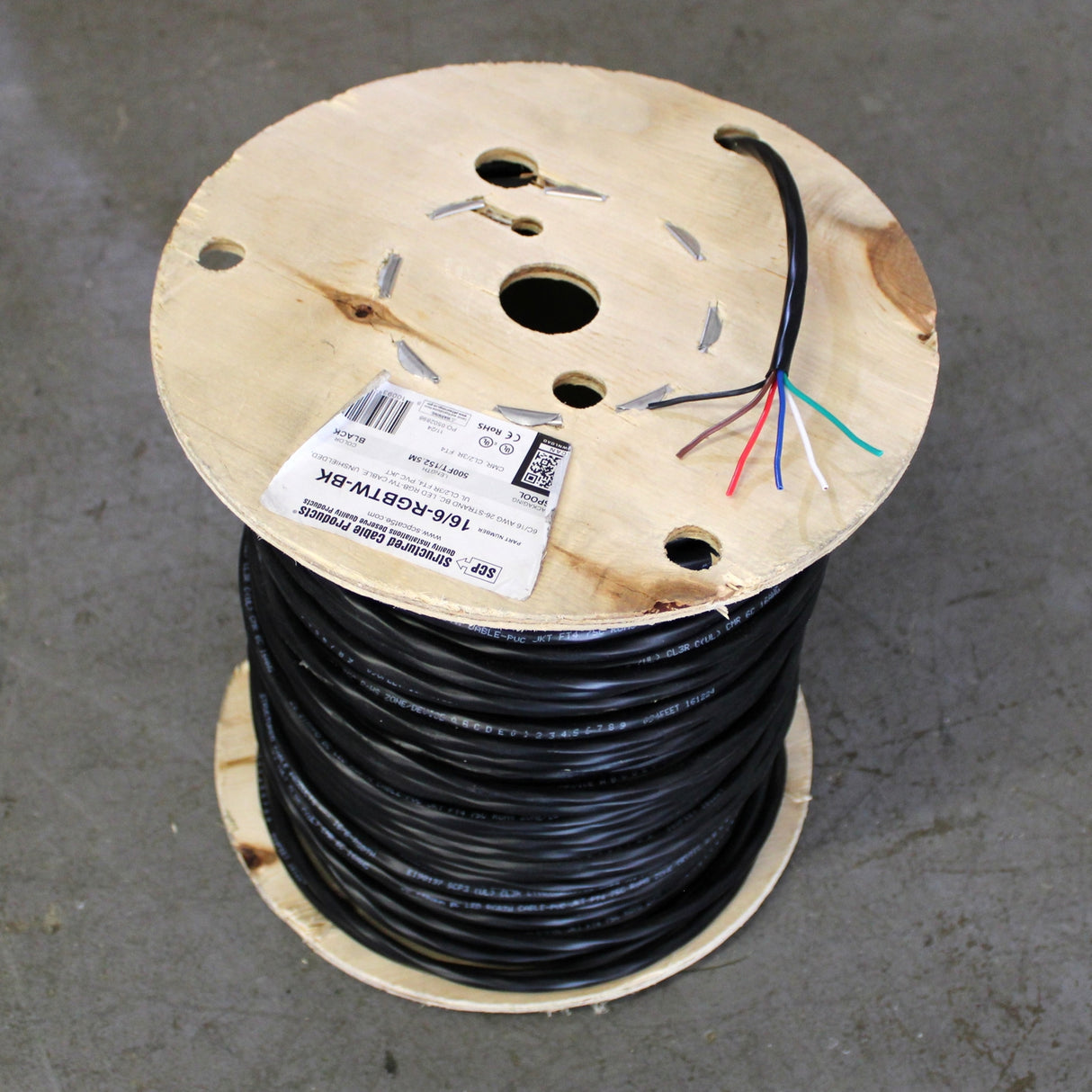 A large wooden spool holds a coil of RGB+TW 6-Core PVC Coated Wire with several colored wires exposed at the top. The labeled spool rests on a gray concrete floor.