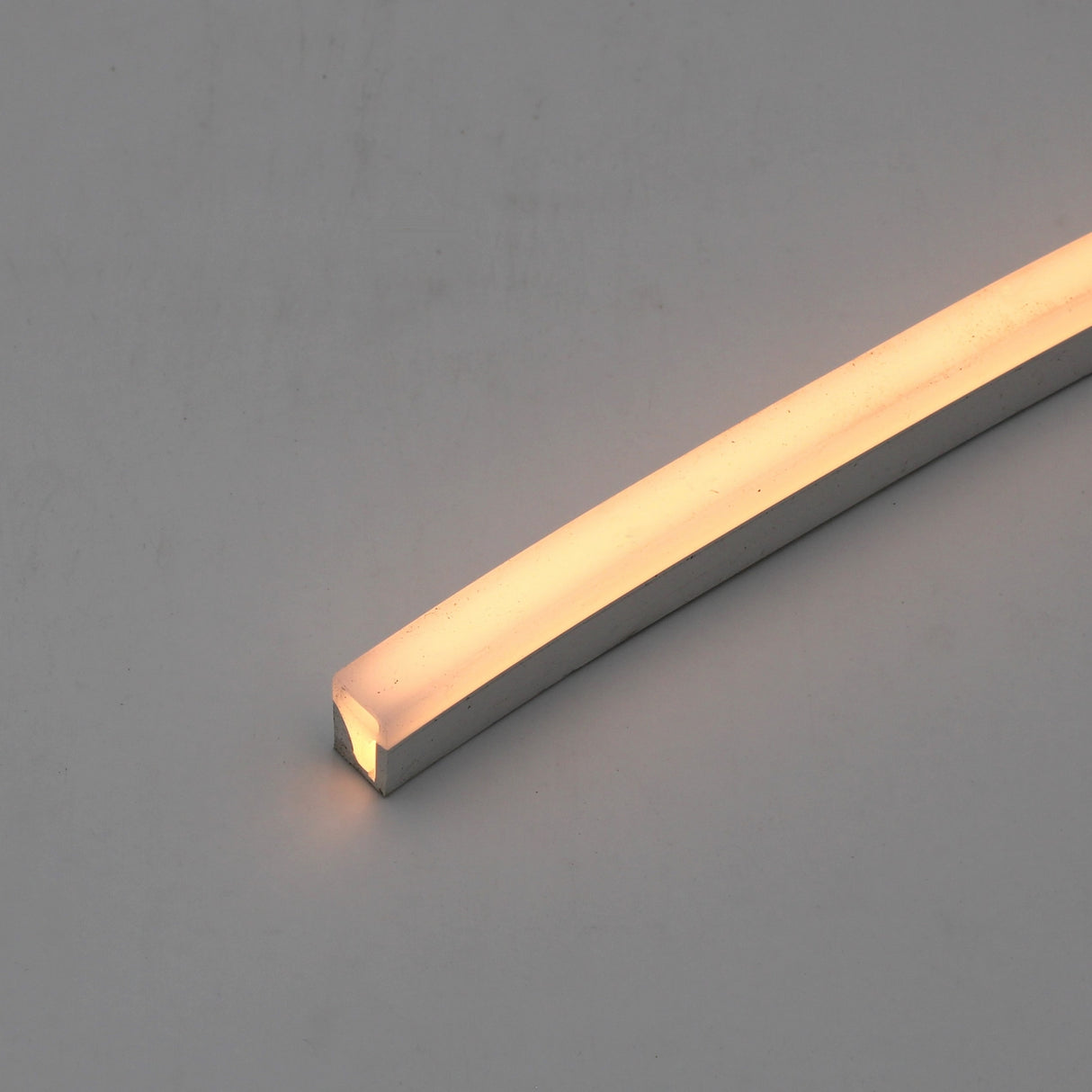 The Flexible LED Neon Light Silicone Profile ~ Model Neon Las Vegas emits a soft warm white glow, ideal for decorative lighting, and is displayed diagonally across a smooth, light gray surface.