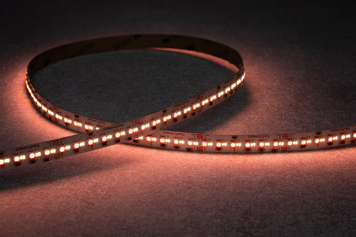 The RGB+CCT IP20 LED Strip (24V) ~ Optimum Cromatic Series emits a warm orange glow in a loose loop on a dark gray textured surface, with evenly spaced bulbs ensuring energy efficiency.