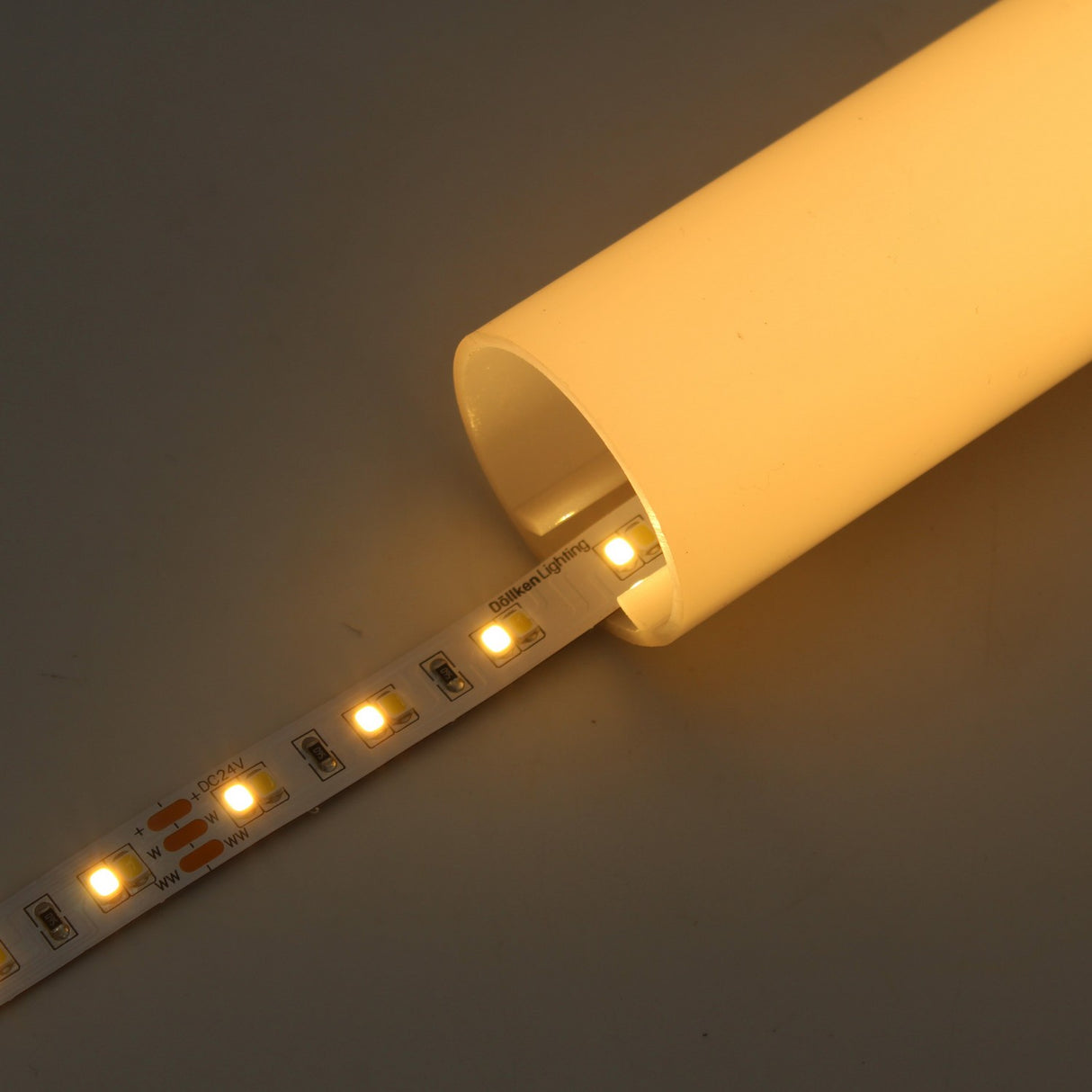 The High-CRI Tunable White IP20 LED Strip (24V) – Optimum Harmony Series emits adjustable warm yellow light onto a neutral surface when partially inserted into a cylindrical, opaque tube, allowing flexible color temperature control.