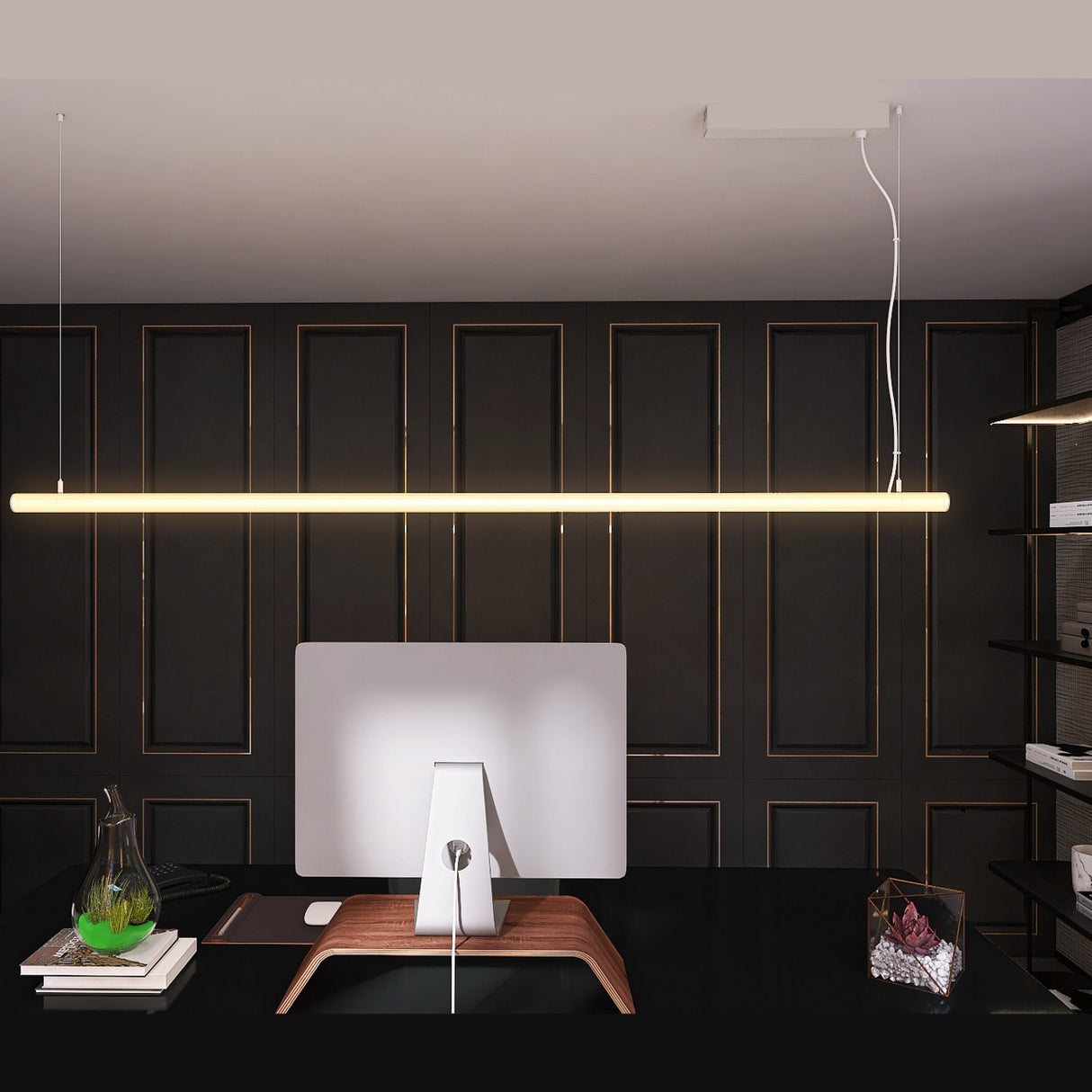 A modern office with a dark paneled wall features a sleek desk, monitor on a wooden stand, books, glass terrarium, and a Cascade38 1.50 Round Acrylic Outdoor LED Lighting Tube with diffuser hanging above the minimalist setup.