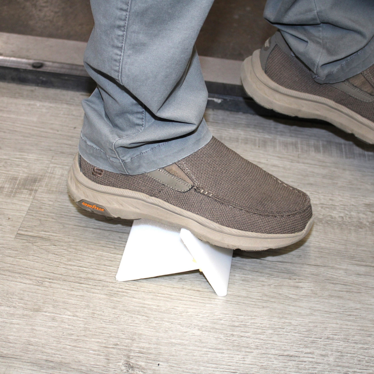 A person wearing gray slip-on shoes and gray pants steps on a small white triangular object—possibly a piece of 2-Part MMA Structural Adhesive for LSE Plastics ~ PE-Bond Kayak500 (50ml 1:1 / B-System)—placed on a wood-patterned floor.