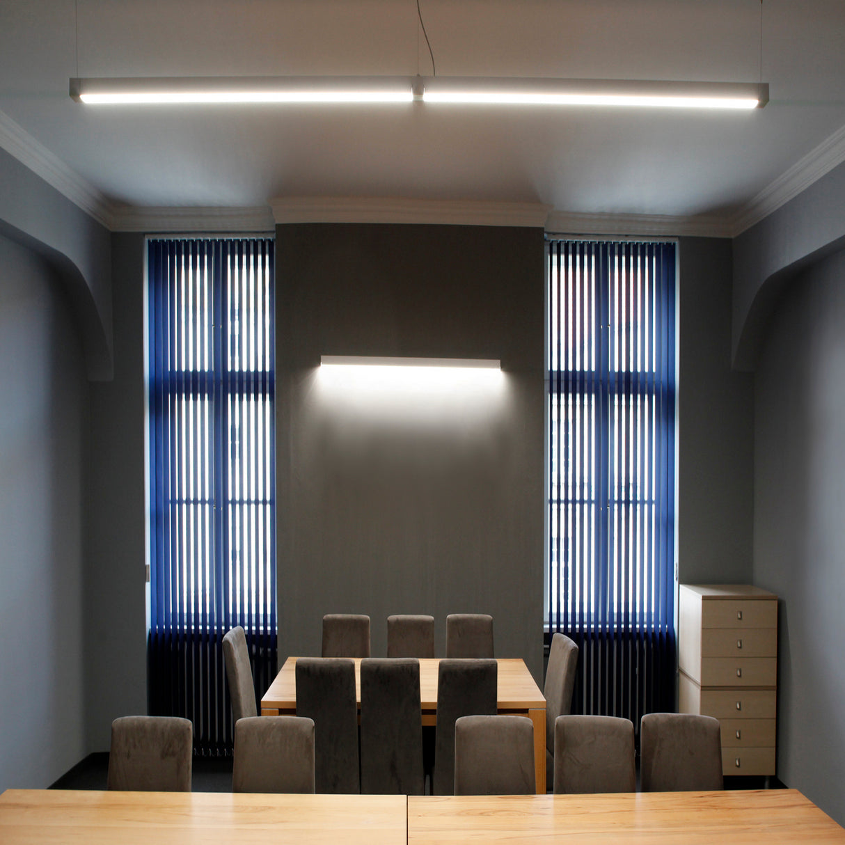 A modern conference room with a long wooden table, gray upholstered chairs, windows with vertical blinds, a filing cabinet, and the 2 Suspended/Surface Mount Ceiling LED Strip Channel ~ Model Phil.v2 creates an inviting professional space.
