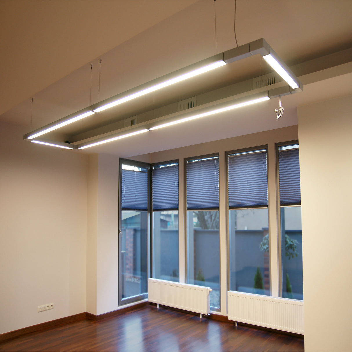 Modern office space with large windows covered by blue blinds, wooden floors, beige walls, and two Phil.v2 2 Suspended/Surface Mount Ceiling LED Strip Channels installed above for stylish commercial lighting.