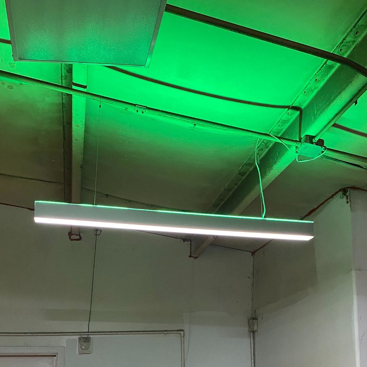A Linear Suspension Lighting LED Direct/Indirect Profile ~ Model DPL55-FL fixture hangs from an exposed ceiling with green lighting and visible beams and wires, creating an industrial-style indoor atmosphere.