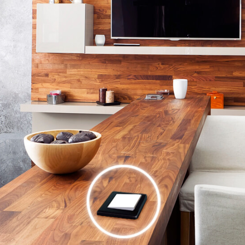 A modern kitchen with a wooden countertop features a wooden bowl of plums, a white cup, kitchen items in the background, and the Push Button Dimmer Switch Wi-Fi Insert ~ inBox by BleBox with a white button on the counter.