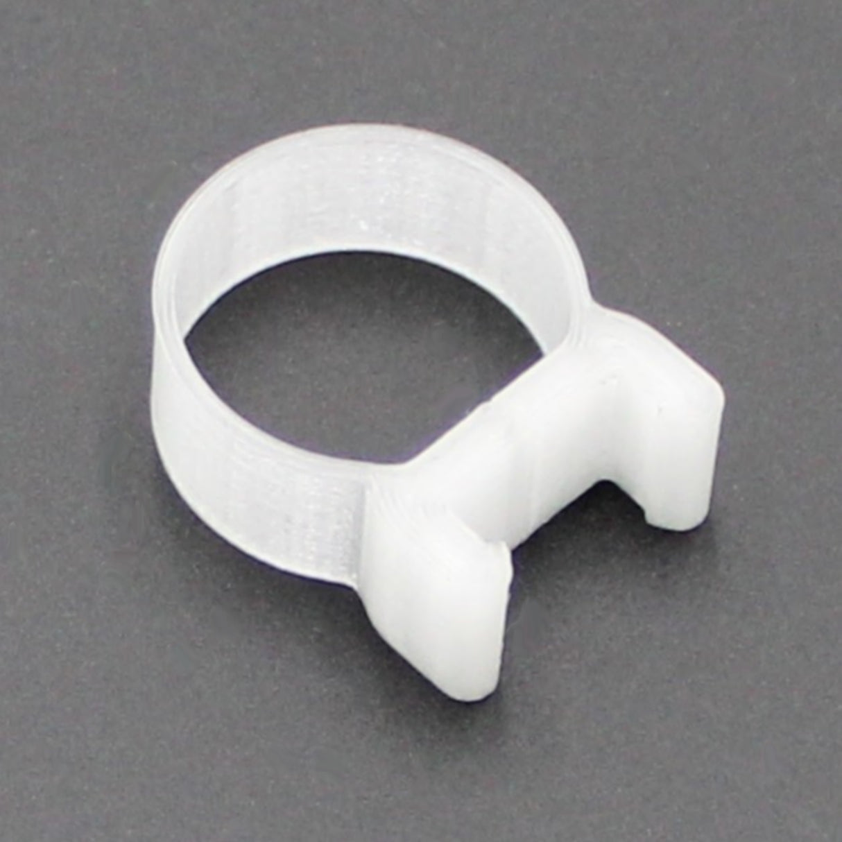 The T-Clip for LED Diffuser Redondo XL is a white plastic clamp with a circular band and two-pronged lock, shown on a dark gray surface, ideal for securing hoses to a T-Section Aluminum Bar.