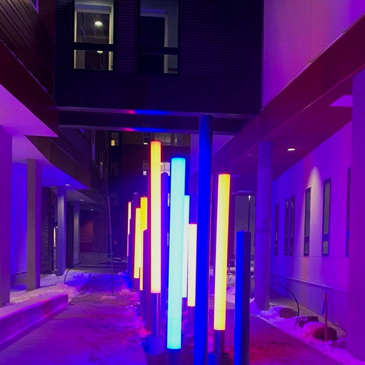 The RGBW LED Pillar Tube Light ~ Model Colorado glows in blue, yellow, orange, and red at night, illuminating a snowy modern outdoor walkway with vibrant colors. Blue and purple ambient lighting enhances the vivid effects between buildings.